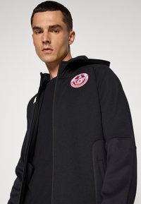 Juodas zip-up džemperis su logotipu kairėje krūtinės pusėje, tekstūruotais rankovių elementais ir suveriamu gaubtu. Lygaus audinio, prigludusio dizaino.