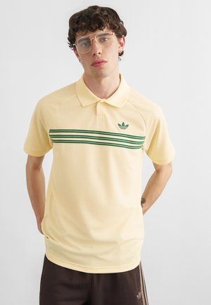 Mladý muž s kučeravými vlasmi a okuliarmi, oblečený do bledožltého Adidas polokošele so zelenými pruhmi a tmavohnedých Adidas nohavíc, stojaci s rukami za chrbtom.