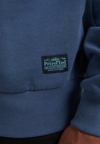 Felpa blu navy con polsini e orlo a coste. Presenta una toppa nera con testo teal che recita "Petrol Ind 1989 Driven by Heritage."