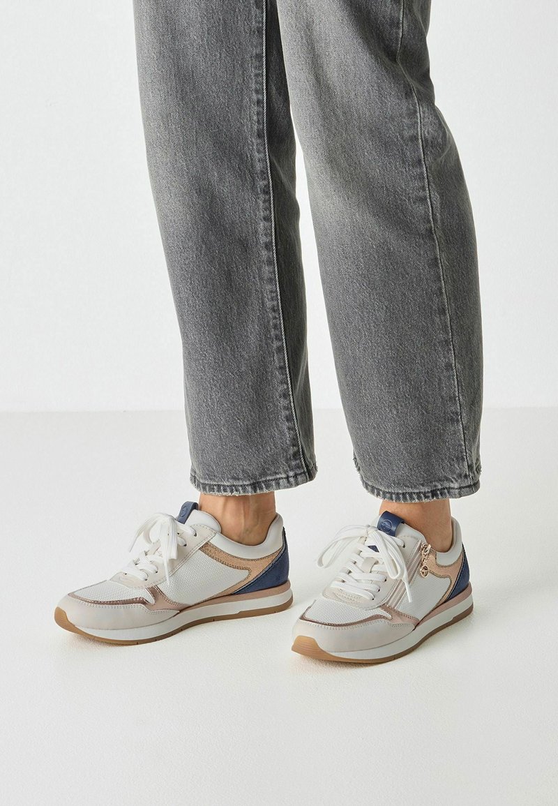 Baskets blanches avec accents bleus et beiges, présentant une tige en mesh texturé, une semelle en caoutchouc et des lacets plats. Portées avec un jean gris court.