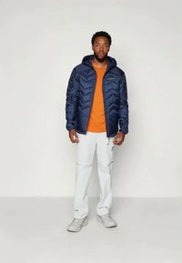 Moški stoji obrnjen naprej, oblečen v modro puffersko jakno, oranžno majico, bele cargo hlače in svetlo siva športna obutev, proti enotnemu ozadju.