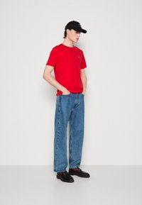 T-shirt rouge à manches courtes, jeans en denim bleu amples et chaussures noires. La casquette est noire avec un logo, portée de côté. Tenue simple et décontractée.
