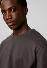 Jeune homme à la peau foncée portant un sweat-shirt gris foncé Calvin Klein, regardant calmement vers la caméra sur un fond gris uni.