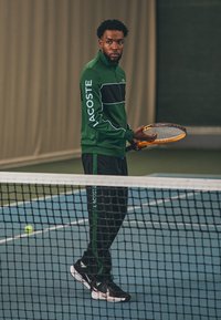 Groene Lacoste zip-up jack met zwarte streep, gecombineerd met zwarte broek met groene accenten. Speler die een tennisracket vasthoudt op een baan.
