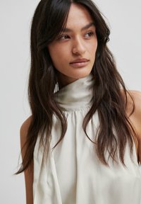 Kremowa bezrękawnikowa bluzka z wysokim kołnierzykiem, wykonana z miękkiego materiału i delikatnie opadająca wokół szyi, tworząca gładki, wyrafinowany wygląd.