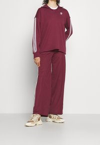 Chándal burdeos con cuello con cremallera y acentos de tres rayas blancas en las mangas y los lados. Presenta una parte superior y pantalones de corte holgado. Zapatillas beige.