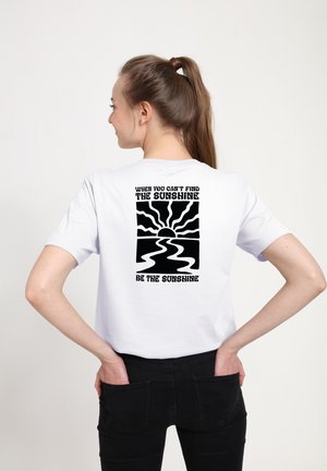 Kvinde i hvid t-shirt med sort sol- og flodgrafik og tekst: "When you can't find the sunshine, be the sunshine."