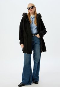 Sort parka med pelsforet hætte, parret med en lyseblå skjorte og højtaljede, brede blå jeans. Sorte solbriller og sko.