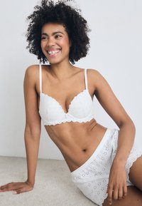Soutien-gorge en dentelle blanche avec des bords festonnés et des motifs floraux, associé à un short blanc en broderie anglaise avec un ourlet volanté. Tissu texturé sur l'ensemble.