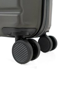 Primer plano de la esquina de una maleta rígida negra que muestra una superficie texturizada y ruedas dobles giratorias con patrón de fibra de carbono.