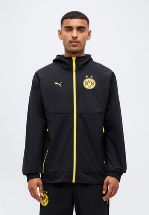 Mężczyzna ubrany w czarno-żółtą kurtkę Borussia Dortmund i pasujące spodnie stoi na jednolitym jasnym tle.