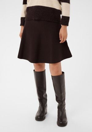 Mulher vestindo uma saia preta evasê, botas de couro pretas até ao joelho e um suéter bege e preto às riscas, em pé contra um fundo simples.