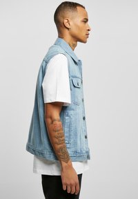 Vest din denim de culoare albastră deschisă, cu guler, nasturi frontali și două buzunare pe piept, purtat peste un tricou alb și pantaloni negri.