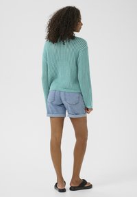 Maglione turchese lavorato a maglia con vestibilità ampia e trama aperta e testurizzata. Abbinato a pantaloni corti in denim chiaro e sandali neri a infradito.