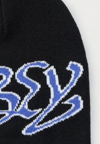 Căciulă neagră tricotată, cu litere albastre și albe îndrăznețe, stilizate. Textură moale, cu o potrivire strânsă și partea superioară rotunjită.