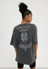 Žena s kovrčavom punđom nosi preveliku tamnosivu košulju s natpisom "Mistično energija" i cvjetnim motivom moljca na leđima, u kombinaciji s crnim kratkim hlačama.