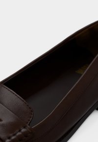 Σκούρο καφέ δερμάτινο μοκασίνι με ραφές στο μπροστινό μέρος και λεία εσωτερική επένδυση, σε απλό λευκό φόντο.