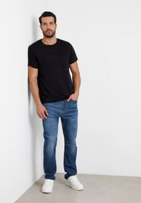 T-shirt nera in cotone con logo "GUESS" in rilievo, abbinata a jeans di denim blu e sneakers bianche. Sfondo semplice, modello appoggiato contro il muro.