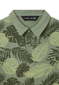 Polo verde con colletto abbottonato, caratterizzato da un motivo di foglie monocromatiche in verde chiaro e verde scuro su uno sfondo testurizzato.