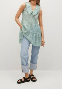 Vaaleansininen kukkakuosisella ilmanhihaisella tunikalla, jossa on rypytetty kaulus, yhdistettynä vaaleanpesuisiin, käännettyjalkaisiin farkkuihin ja tukeviin mustiin sandaaleihin.