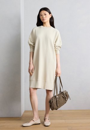 Kvinde iført en beige langærmet sweatshirtkjole, beige flade sko, holder en brun rynket håndtaske, stående på et trægulv foran en grå væg.