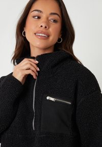 Sort fleece pullover med tekstureret stof, høj krave, halvmatchende lynlås foran og en lynlåslomme på brystet, der er fremhævet med sort materiale.