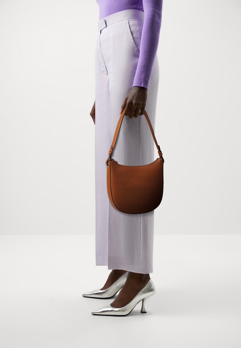 Bolso de hombro de cuero marrón claro con diseño perforado, sostenido por una persona que lleva pantalones de pierna ancha de lavanda clara y tacones de punta metálica plateada.