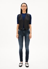 Svart ärmlös väst med knappar framtill, bärs över en marinblå ribbad skjorta och matchas med mörka slim-fit jeans samt svarta loafers med vita detaljer.