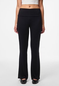 Vrouw die zwarte, rechte, high-waisted broek en zwarte ballerina's draagt, gecombineerd met een wit crop top, staande tegen een witte achtergrond.