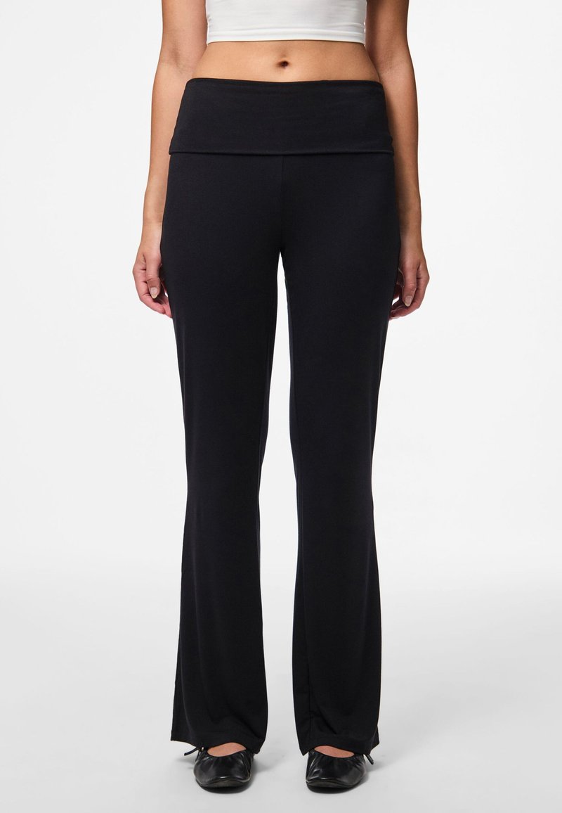 Vrouw die zwarte, rechte, high-waisted broek en zwarte ballerina's draagt, gecombineerd met een wit crop top, staande tegen een witte achtergrond.