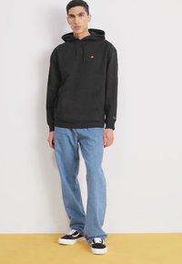 Sudadera con capucha negra, jeans holgados azules y zapatillas negras con suelas blancas. Detalle de logo notable en la sudadera.