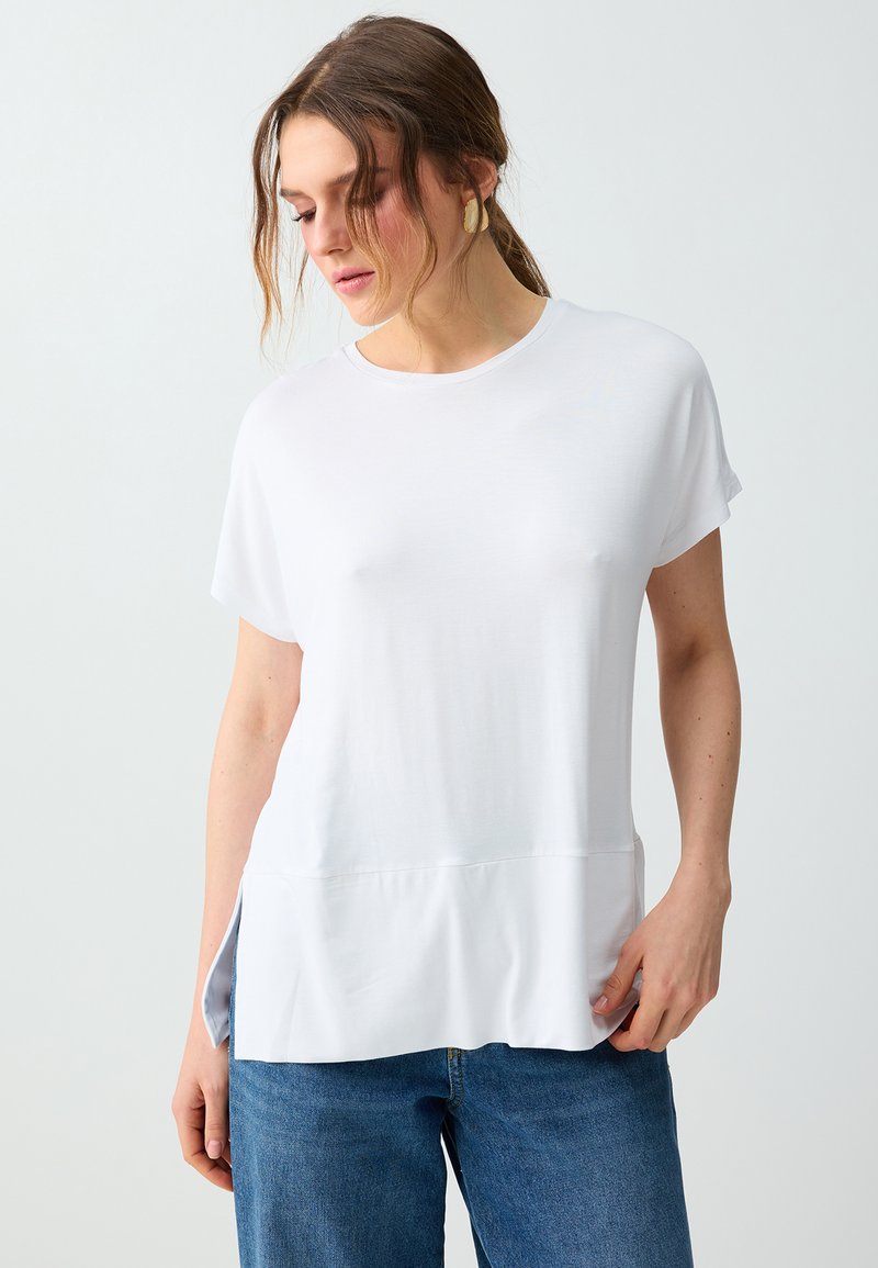 Femme à la peau claire et aux cheveux bruns porte des boucles d'oreilles en or, une chemise blanche ample à manches courtes avec des fentes sur les côtés, et un jean bleu sur un fond uni.