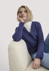 Lila V-Ausschnitt Pullover aus Strick mit gerippter Textur, getragen über einem weißen Rollkragenpullover, kombiniert mit hellblauen Jeans. Vor einem neutralen Hintergrund.