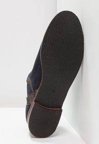 Suela de un zapato azul oscuro con una suela y tacón de goma negra texturizada, talla 37, posicionada verticalmente contra una pared blanca.