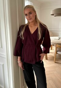 Top burgund cu decolteu în V, mâneci largi și talie legată, purtat împreună cu pantaloni negri cu talie înaltă. Amplasat într-o cameră modernă, luminoasă.