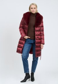 Bordeaux puffer coat met een nepbont kraag, met een glanzende afwerking, horizontale stiksels en twee voorkruiken, gedragen over een bruine turtleneck.