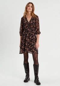 Robe fleurie noire avec un décolleté en V, des manches trois-quarts, une taille froncée et un tissu transparent ; portée avec des bottes noires arrivant au genou.