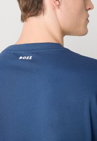 Marineblå bomulds T-shirt med rund halsudskæring. Har hvid "BOSS" logo på bagsiden nær kraven. Glat tekstur og simpelt design.