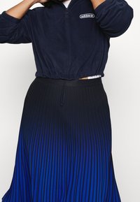 Person som bär en marinblå Adidas fleecekortad jacka och en plisserad kjol med ett svart-till-blått gradientmönster.