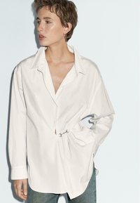 White button-up shirt made of cotton, featuring a loose fit, oversized collar, large cuffs, and a gold accent near the waist.