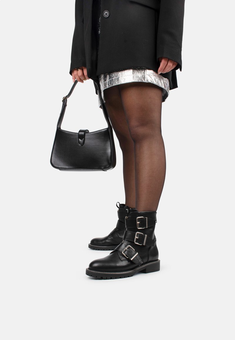 Botas negras de cuero hasta el tobillo con múltiples hebillas plateadas, suela texturizada, usadas con medias negras transparentes y una falda plateada, complementadas con un bolso negro.