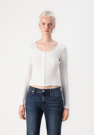 Hvit, langermet, ribbestrikket crop-top med rund hals og dekorativ knappestolpe. Kombinert med mørkeblå jeans.