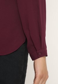 Camicia bordeaux a maniche lunghe con una texture morbida, dotata di polsini abbottonati e orlo curvo. Indossata sopra un tessuto scuro.