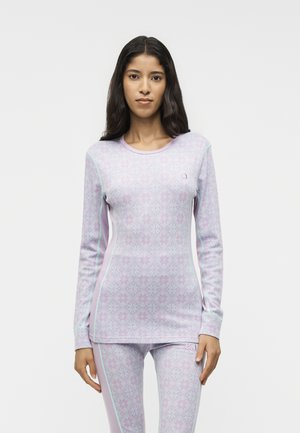 Vrouw met lang donker haar die een nauwsluitend lavendelpatroon langemouwen topje en bijpassende legging draagt tegen een effen witte achtergrond.