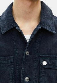 Kostymjacka i mörk marinblå färg, med klassisk krage, silverfärgade tryckknappar och två framfickor med distinkt horisontell ribbstruktur.