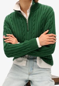 Vihreä neulottu neule, jossa on palmikkokuvio, puolivetoketjullinen kaulus ja resorit hihansuissa, käytetty valkoisen kauluspaidan päällä. Siinä on pieni Amerikan lipun tunniste.