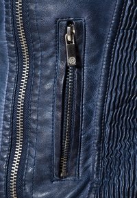 Giacca di pelle blu marina con superficie testurizzata, chiusura con_zip in argento e linguetta rettangolare, e dettagli di cucitura a coste.