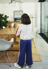 Pull en tricot blanc avec une rayure rouge, associé à un pantalon large bleu avec des boutons blancs sur le côté. Ambiance intérieure aux tons neutres.