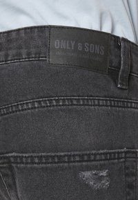 Vaqueros de mezclilla negros con una etiqueta de tela con el texto "ONLY & SONS". Detalles desgastados en el bolsillo trasero. Diseño clásico de cinco bolsillos.