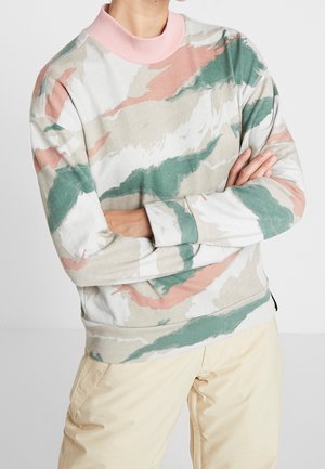 Person trägt ein Sweatshirt mit Rundhalsausschnitt und langen Ärmeln mit grünen, beigen und rosa abstrakten Mustern und cremefarbenen Hosen, die Arme verschränkt.